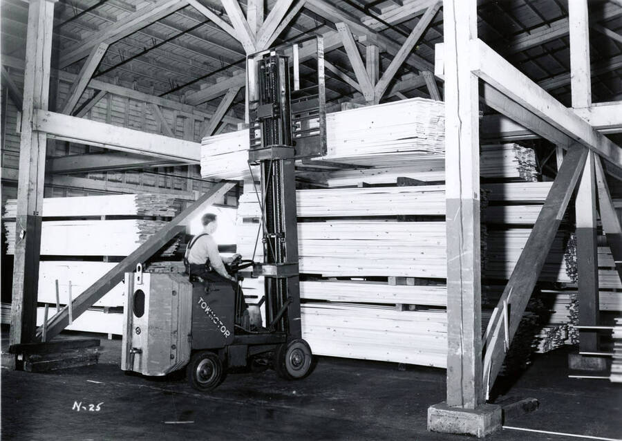 A man uses a forklift to lift a stack of lumber on to a rack. The description on the back reads "Lumber carrier,  #24 pressed shed."