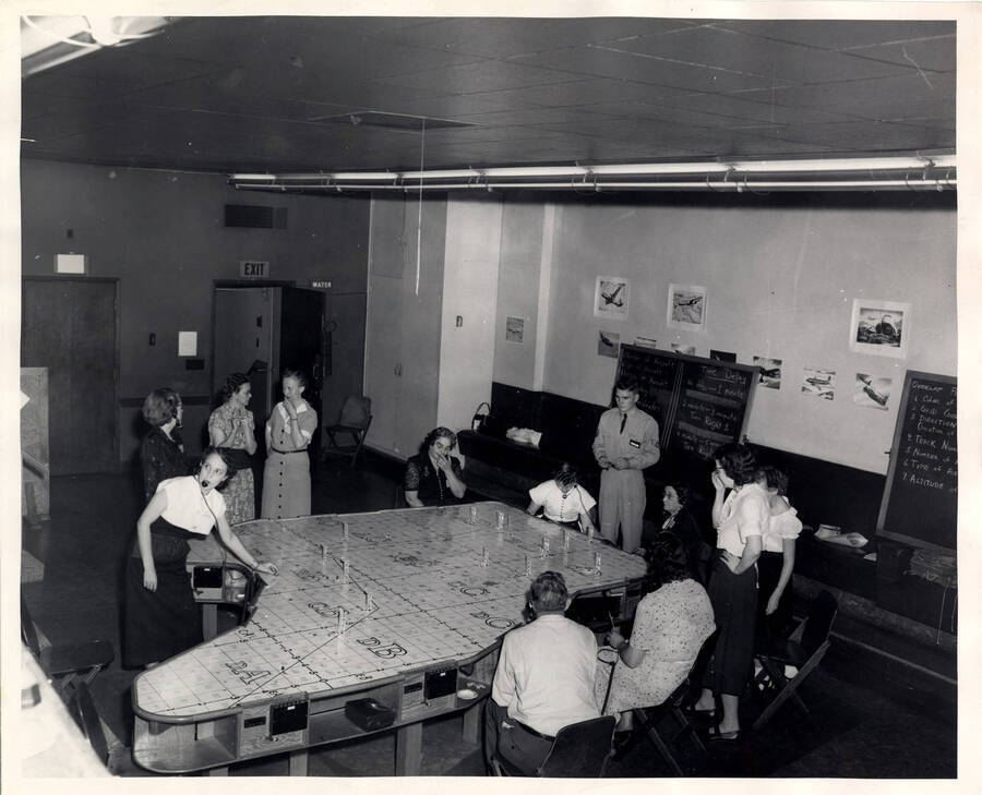 The description on the back of the photograph reads "Operations room in the Spokane filter Center. Civilian volunteers plotting aircraft." Men and women stand around a lot plot map.