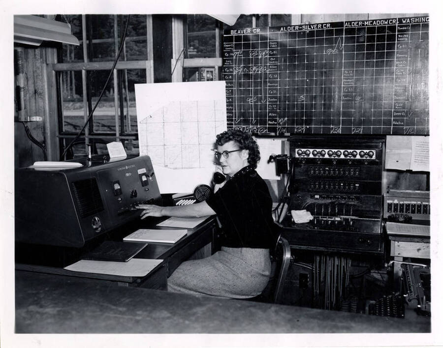 Cora Todd uses the radio in Lewiston, Idaho. Behind and to her right is the locomotive schedule for the lumber trains.