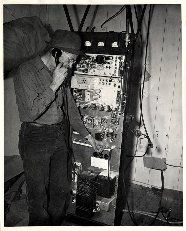 The description on the back of the photograph reads 'Forrest Vaughan checks transmitting equipment in shack on Bald Mountain, where one of two transmitting towers which controls PFI radio systems is situated.'