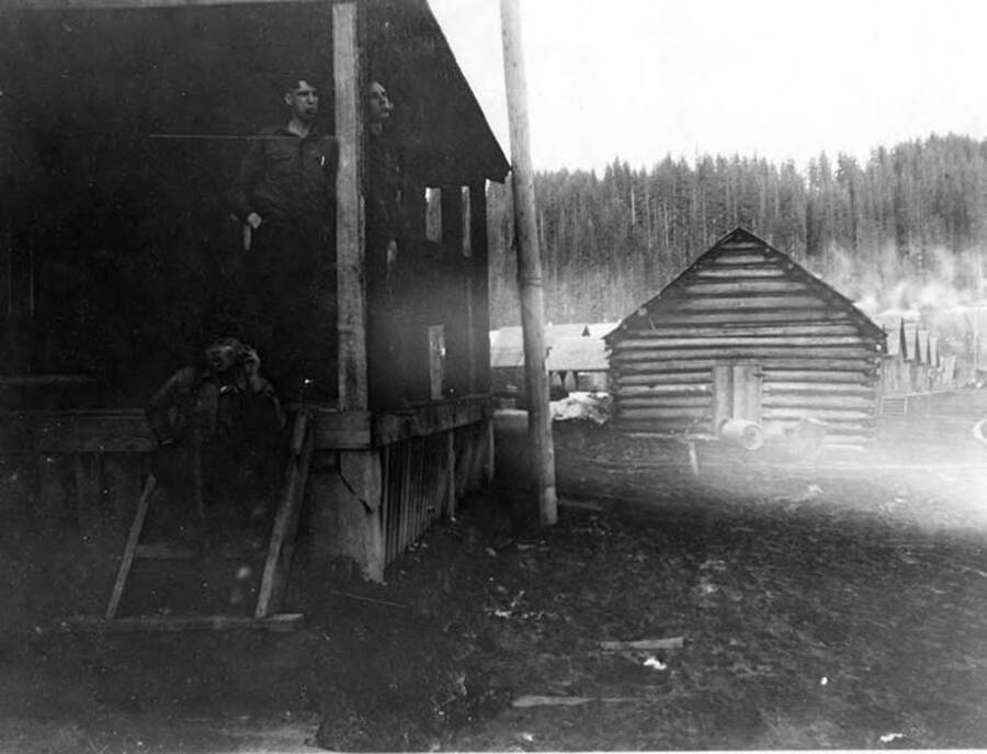 Three men sit on the porch of the store building in Headquarters, ID. The description on the back of the photograph says that the log building in the background area is now occupied by Headquarters Dry Store.