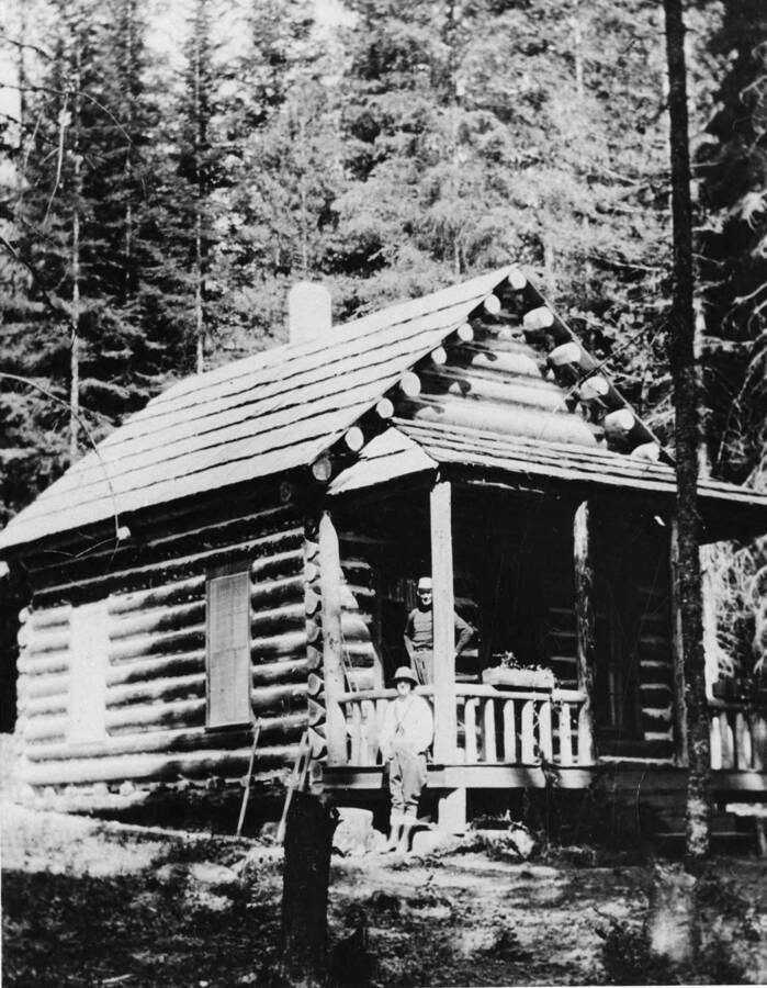 A man stands on the porch while another stands below him on the ground at a log cabin in the woods. Written on the back of the photograph is 'reprinted from old Beat Curtis photo. Copy negative made August 1958.