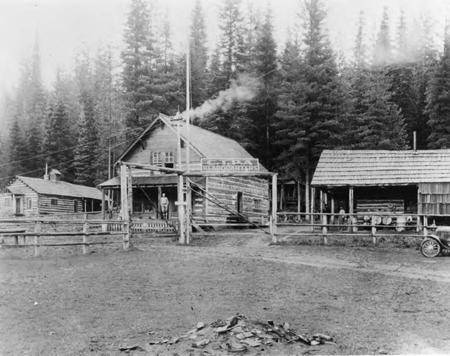 In the foreground, a sign reads C.T.P.A Headquarters (Clearwater Timber Protective Association). In the background, a man leans against a post of a log building. On the envelope holding the picture: Copy negative of the original (first) Clearwater Timber Protective Assn. Headquarters dated 8-26-26. This building still stands on this site at the Potlatch town of Headquarters. The building was used by Potlatch after the CPTPA moved to it's present site on "White's Meadow" for a transient's bunkhouse, then as a grocery store and most recently (as of 1987) as a saw shop.