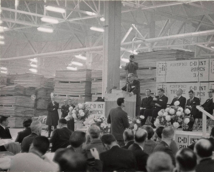 Dedication of the Jaype Mill. A man is standing at the podium speaking with other officials seated behind him. A man stands behind him photographing the scene. Boards of lumber are also seen behind him.