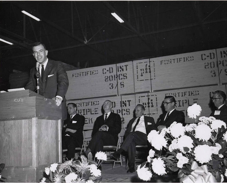 A speaker speaks at the Jaypee Mill dedication. Behind the speaker are other speakers as well as stacks of wood stamped and read for distribution.