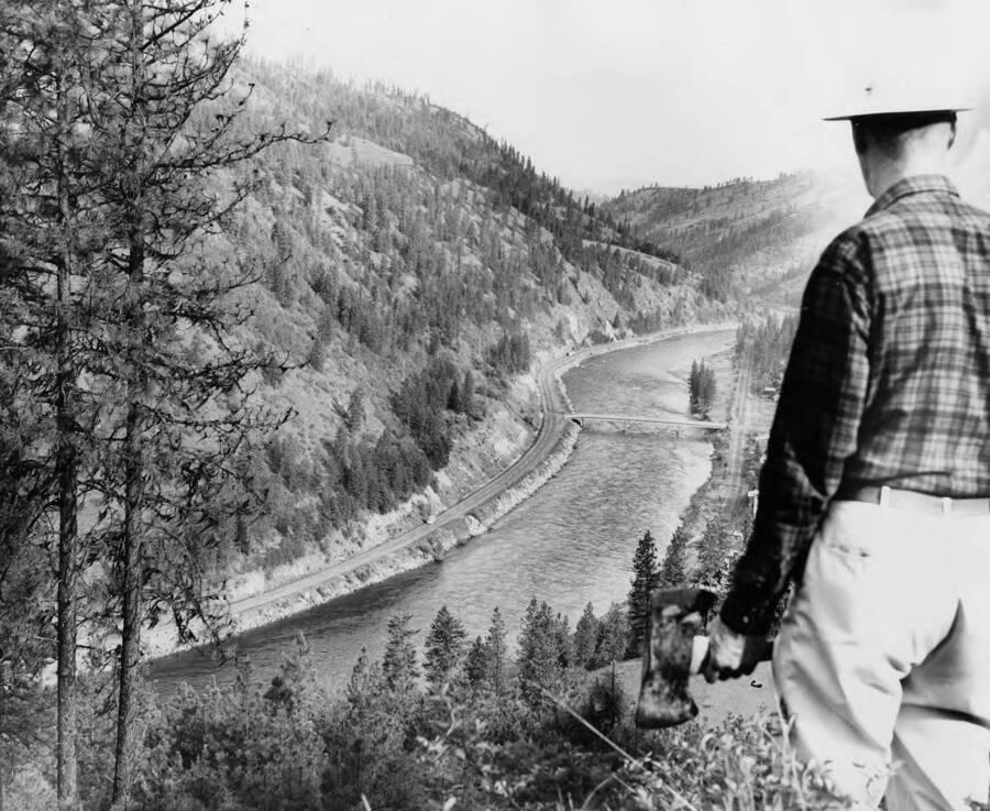 A man holding an ax stands on Greer Grade and the town of Greer, Idaho. The description on the back of the photograph reads 'traveling to the headquarters of the Clearwater log drive, one leaves U.S. Highway 12. U.S. Highway 12, curing along the far side of the Clearwater river in central Idaho, brings, one to within fifty miles of Camp T. Headquarters of Potlatch Forests, Inc. log drive, last in the nation. This viewpoint on the steep and winding Greer Grade looks down on the hamlet of Greer, clustered between forest and river.'