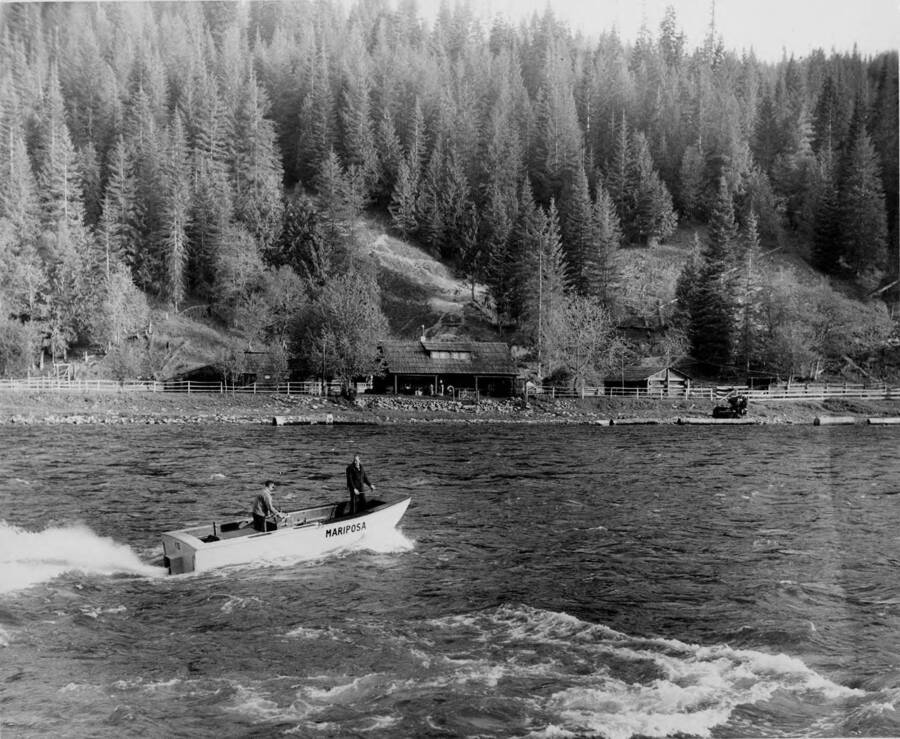 The jet boat "Mariposa" powers across the river. On the bank of the river is a farm as well as logs.