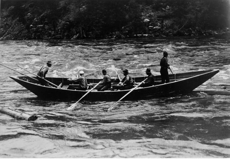 Potlatch men use oars to propel a boat through the Clearwater river in a log drive. Attached to this photograph is a paragraph that reads 'backbreaking stamina was the order of the day for these Potlatch oarsmen several decades ago, when all they had to fight the turbulent Clearwater river with was a heavy wooden bateau and eight sturdy arms. in 1949, the bateau was modified to take a small outboard motor; today's modern boats first appeared on the log drive in 1959'.