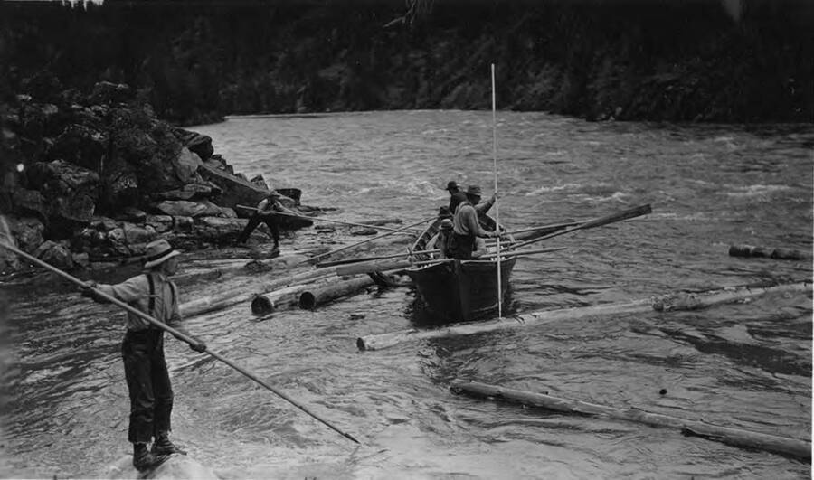 The men in the boat use oars and long poles to pull logs into the Clearwater River while two men on shore use long poles to help. The description on the back of the photograph reads 'bateau crew at work on North Fork 1939.'