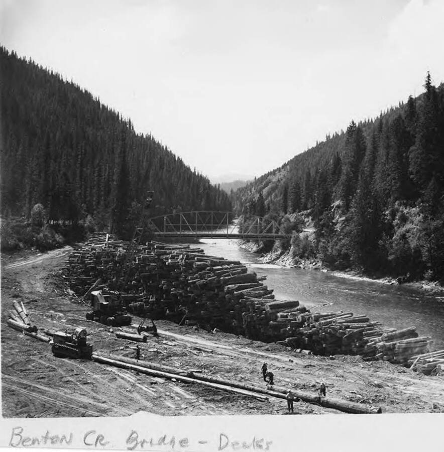 The log deck on Benton Creek in Idaho. In the background is the Benton Creek Bridge. Men work at the bottom of the photograph to prepare the logs to be stacked on the log deck.