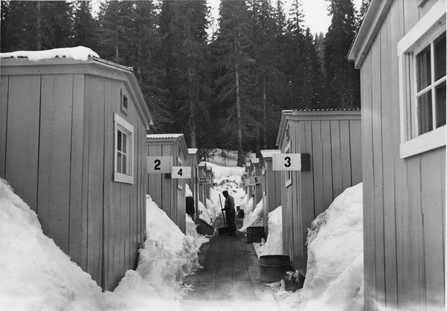 The bunkhouses numbered 2, 3, 4, 5, and the wash house of Camp 44. A man stands in the isle with a broom. The snow is piled up fairly high in-between the buildings.