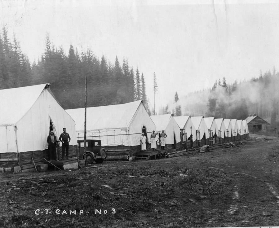Two loggers stand in front of their bunkhouse in camp 3. In the next building over, three men, possibly cooks, stand in front of another bunkhouse. The description on the back says 'About 1 mile east of Hollywood [Idaho] Cook on left is Fred Thomas. Cook in center is Marco. Car is 1927 Chev.'