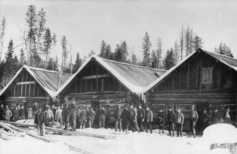 Loggers stand in front of log cabin bunkhouses at a logging camp near Bovill, Idaho.