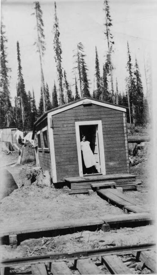 A woman leans in the doorway of a bunkhouse in Camp 6. In front of her is an elevated wooden platform as well as railroad tracks.