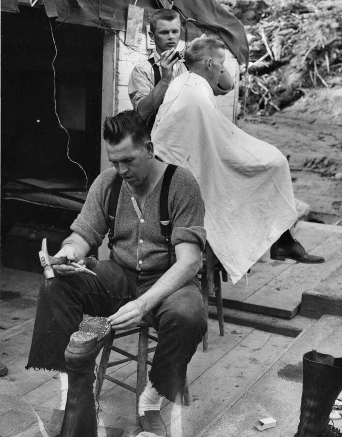 One man works on one of his boots, replacing the metal studs while behind him, another man gets his haircut. Written on the back of the photograph 'cooking up' and 'ear lifting.'