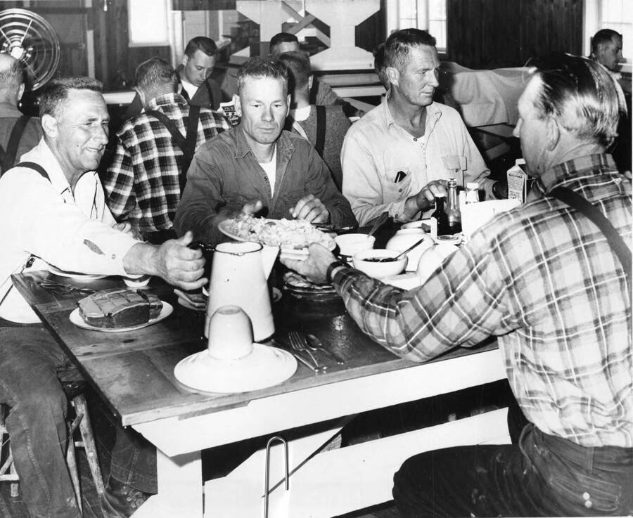 Men dine in the mess hall in Camp T. Attached the photograph is 'hearty breakfasts, complete with scrambled eggs and cake, fuel men for hard work of the log drive. Meal is cookshack-mess hall at Camp T, as men work upriver from Camp, in clean-up operation to break out log jams. County upriver is roadless, so men boat to their day's work site, return to camp, until moving downriver, when they eat on wingan. Silence at meals is old-time tenet of logging camps. Cooks want eaters to clear out soon so he can set up for next meal.'