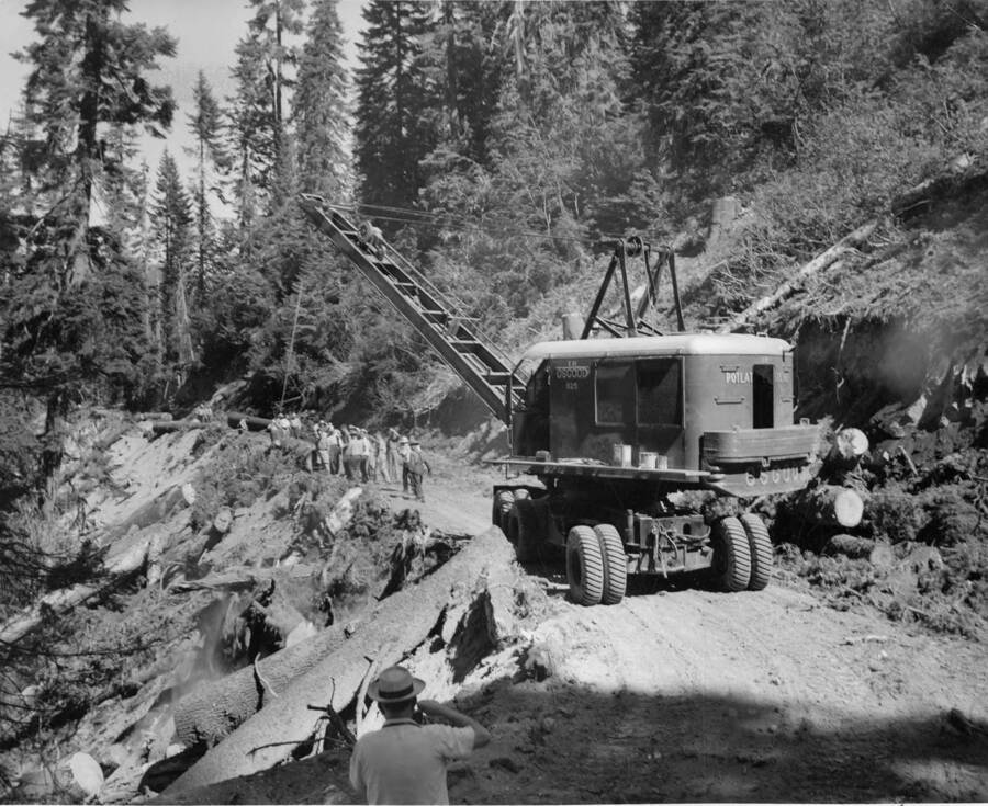 An Osgood loader crane moves up a road. In front it stand a group men, looking over the side of the road at logs.