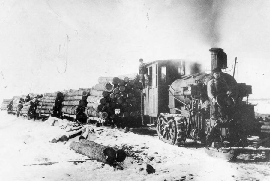 Log sleighs being pulled by a early caterpillar which is then driven by a man on a sled.