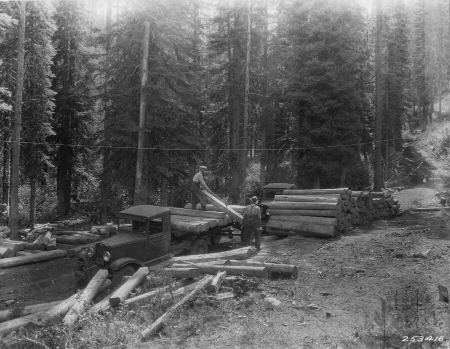 Two men load cut and stripped lumber onto early model trucks.
