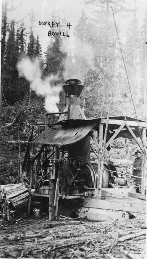 The steam donkey near Bovill, Idaho. This, according to writing on the photograph, is donkey #4. One man standing behind the donkey while another holds the control on the cable wench.