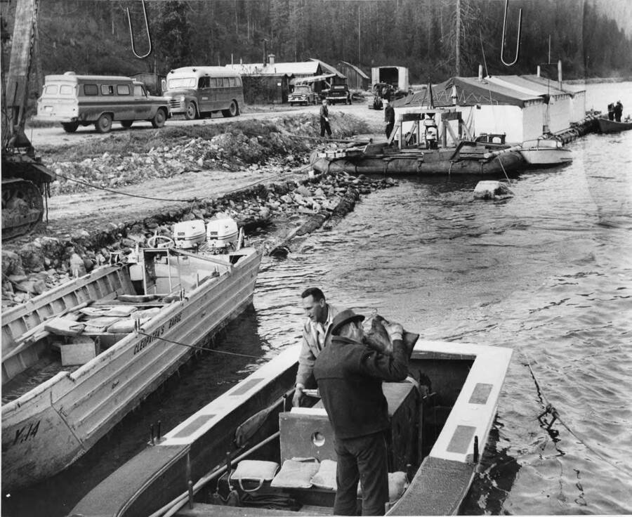 Setting up Camp T in preparation for men working on the log drives. In the background, the floating mess hall and cookhouse, while in the foreground, men prepare the boats. One man drinks from a canvas canteen. Attached to the picture is 'Days at Camp T, log drive headquarters, start early, with men readying jet boats, foreground, that will transport lumberjacks to log jams in river. Drive's wanigan, cook shack, bunkhouses, on three rubber rafts, linked together loosely, to ride the wild rapids is steered by motors at fore and aft, visible on rear raft. men in foreground drinks from canvas canteen.'