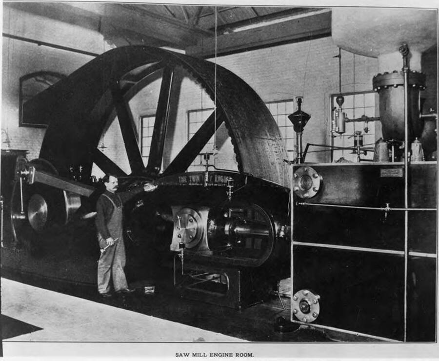 The large engine that drove many machines in the saw mill. A man stands next to it for size comparison.