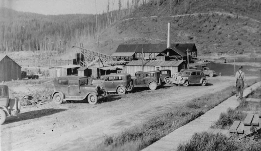 The Lewis mill near Bovill on the East Fork of the Potlatch river. Cars are parked along the dirt road as a man on the right hand side of the photograph walks along the wooden sidewalk.