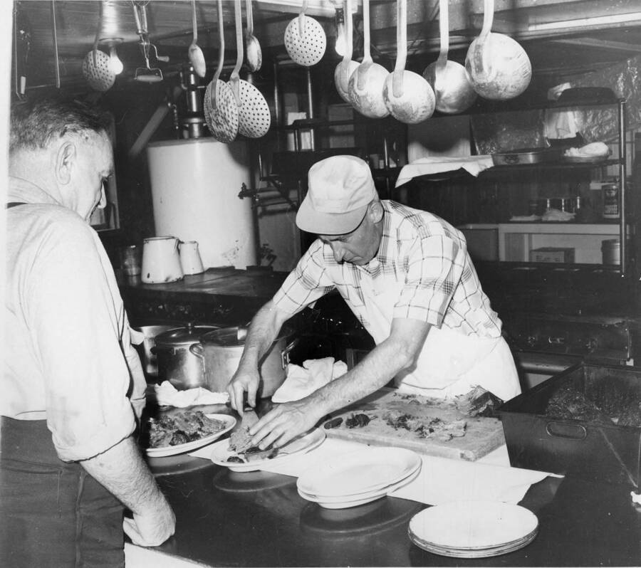 A lumberjack waits for his meal being served by the cook. Attached to the back, it reads 'Ladling out noon dinner of roast beef, Cook Harvey Spears is assisted by flunky 'Michigan' Stoewell, who began log drive work in 1923, now is retired from active log funning, but remains on the scene as cook's helper.