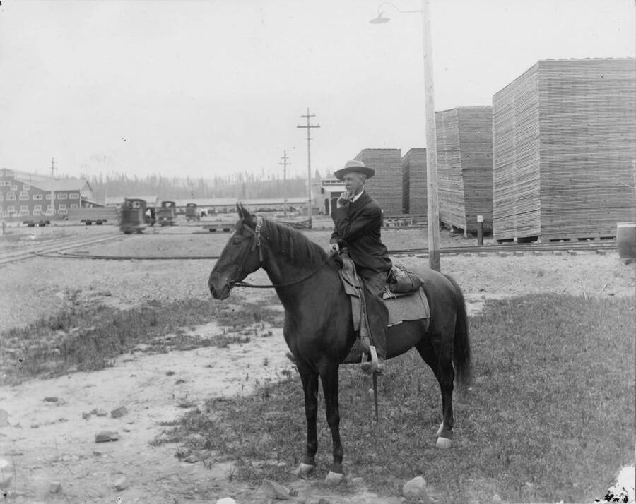Charles Roger sits on horseback in the lumber yard at the mill in Coeur D'Alene. Behind him is the mill as well as stacks of lumber.