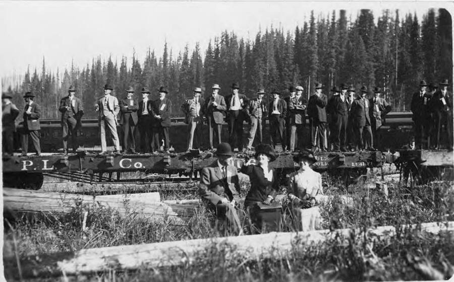 Company men stand on flatcars. Behind them stand unlogged forests. The three sitting on the log are listed from left to right as Mr. Laird, Mrs. Donovan, and Mrs. Knapp.