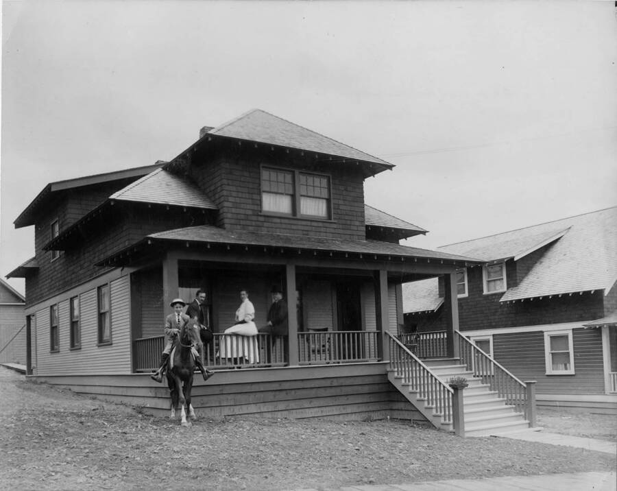 Stanley Smith sits on his horse while behind him, Mr. and Mrs. Smith sit on the railing (Mr. Smith was the first postmaster). Next to Mrs. Smith is James Fiandmen. They are on the porch of a company house in Potlatch, Idaho.