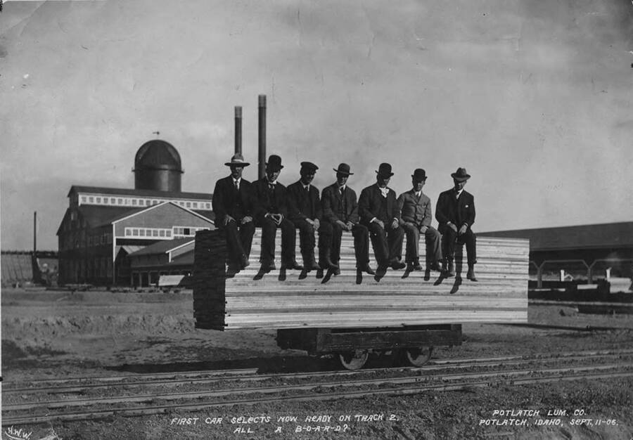 Men of the Potlatch company sit on a load of lumber board. The caption for the photograph reads 'first car selects now ready on track 2. All a B-O-A-R-D?' Behind the men is the Potlatch sawmill in Potlatch, Idaho. From left to right: R.M. Weyerhaeuser, Cliff Musser, William Deary, A.W. Laird, Mr. Wilkinson, A.H. Irving and Mark Seymour.