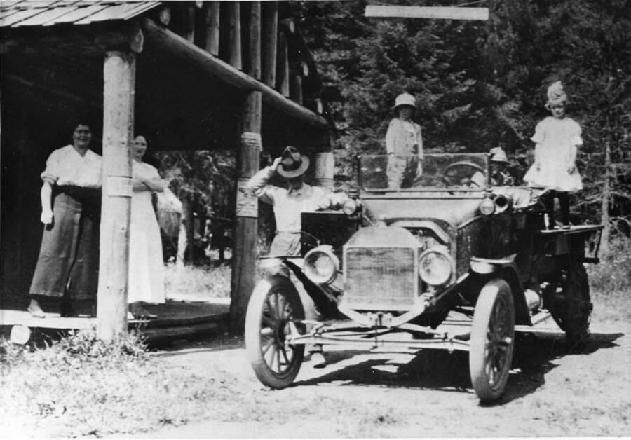 Two women stand on the porch of a log cabin, while two men and two children pose with their car.