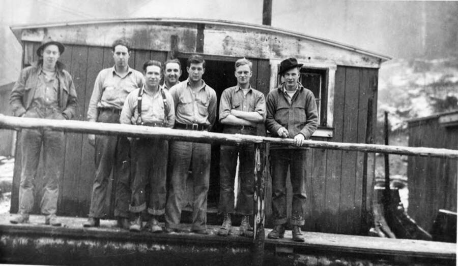 Left to right: unknown, Bighom, A. Hughes, C. Crane, B. Hobbs, B. Shook, and G. Hughes stand on the wooden walkway in front of a bunkhouse.