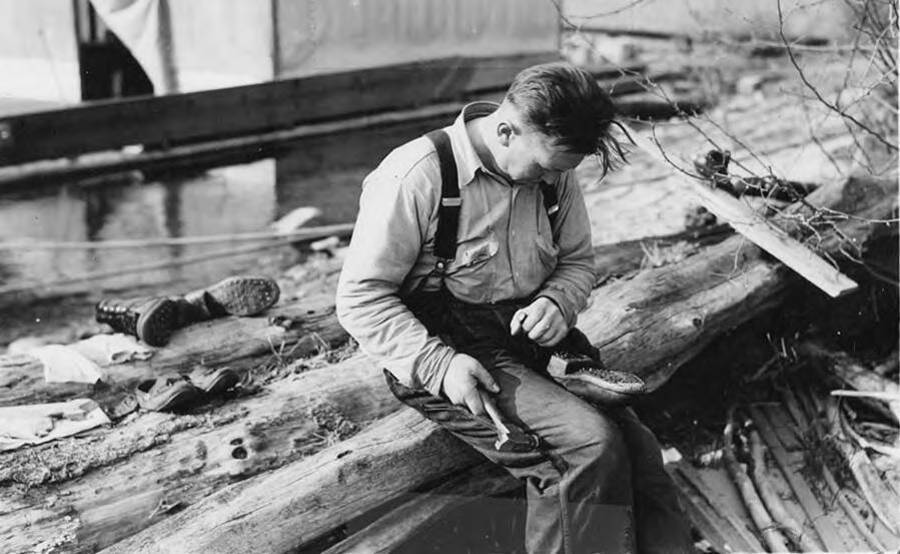 A lumberjack sits near the wanigan repairing his boots. Written on the back of the photograph is 'log drive 15.'