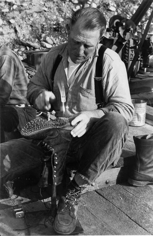 A lumberjack sits and adds new caulks his boots. The description on the back reads 'Dinner over, a Potlatch driver spends a few moments repairing the "corked" (caulked) boots so vital to his safety while working the log jams. Others pass the time reading, playing horseshoes, or spinning a few yars around the campfire.'