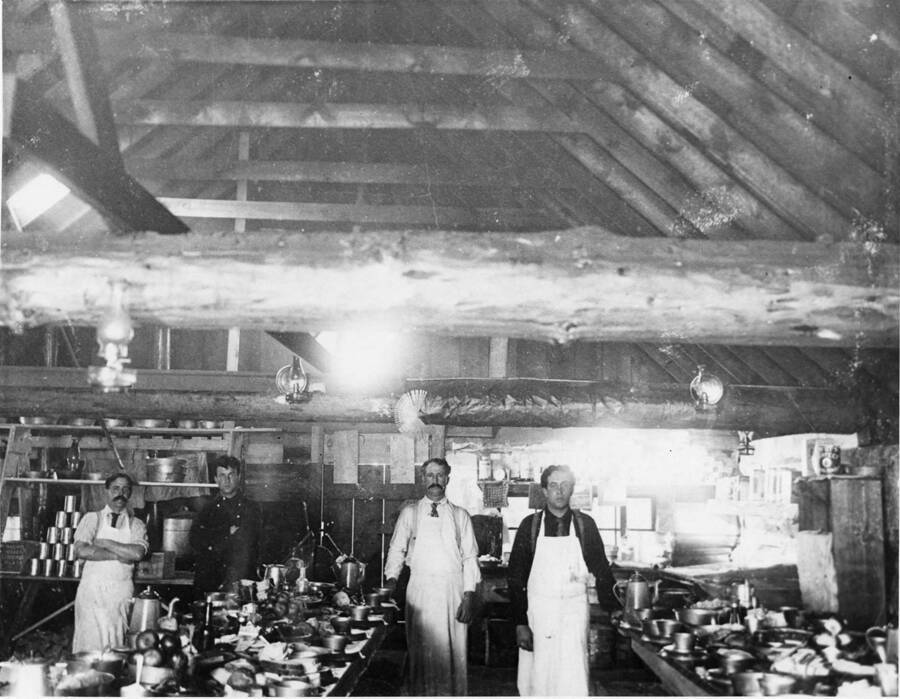 Four man stand in a log cabin serving as the camp cookhouse. On both tables are the dishes used to feed the men of the camp.