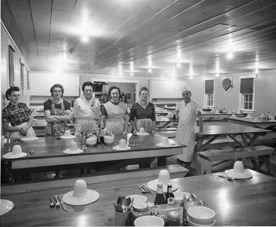 The man and women of a camp kitchen. The women stand in front of a set table with coffee cups and saucers while the man (possibly the cook) leans against an unset table.