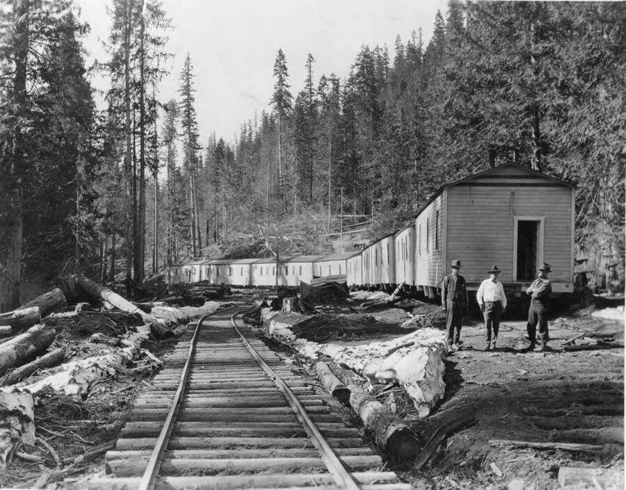 Three men stand to the side of a railroad track. Behind them are railroad bunkhouses.