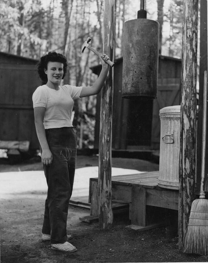 Ms. Adair holds a hammer reading to ring a bell in camp. 'Dolly Adair, attractive flunkey at Camp 27 on Breakfast Creek. Although women flunkeys were used in logging camps during World War I, none were employed at camps in the Clearwater forests until the summer of 1942. (Logging of consequence did not begin in the Clearwater until 1926). Many and dire were the predications of what would happen when the girls went to work in the camps, but aside from a sizeable number of school teacher lumberjack marriages and a noticeable sprucing up by all the men in the camps, nothing much happened. The girl flunkeys have worked efficiently and well. A tendency to move about and to not stay in any one camp for more than a few months sometimes weeks, is evident among them. The beckoning finger of other jobs provides a well nigh [sic] irrestible lure with a resulting high turnover of flunkey labor, but, nevertheless they have performed a fine war job.' Description taken from back of photograph.