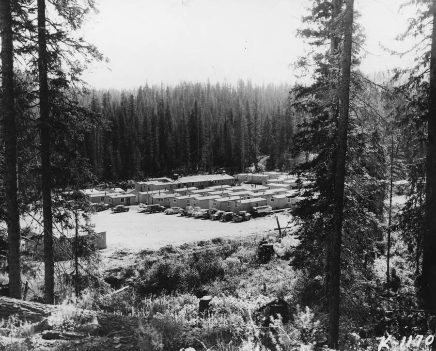 The description on the back of the photograph reads 'Used for stockholders meet 5/15/1958.' The bunkhouses are seen as well as lumberjacks cars and pickups.