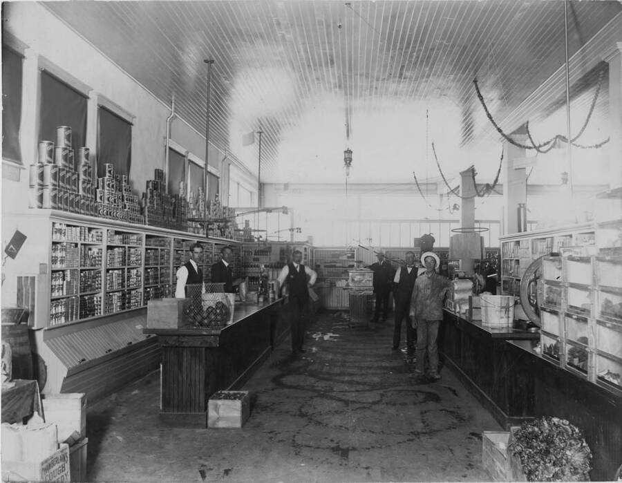 Description from the back of the photograph reads 'This is the present Potlatch Sears Co. store (Grocery dept) in the early days. Along west wall Manning Fansler, in white sleeves behind counter. Charles McDonald in the white hat and Against the counter (moustache) is perhaps Mgr. McDonald.'