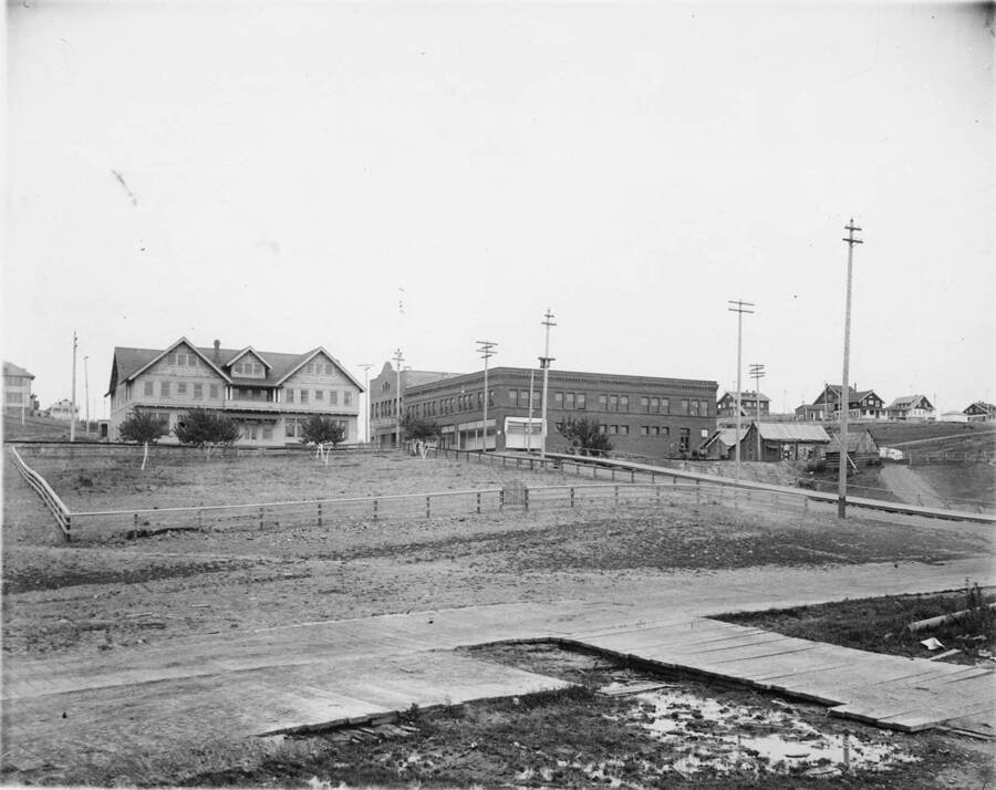 The town plaza of Potlatch, Idaho. In the foreground of the photograph are wooden sidewalks. The house in the background may be a hotel. The building opposite that, may be a store of some kind.