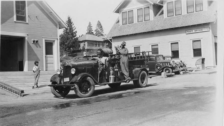 A boy stands watching a fireman shooting water out of a fire house near the post office in Potlatch, Idaho. Another man sits in the driver's seat of the fire truck.