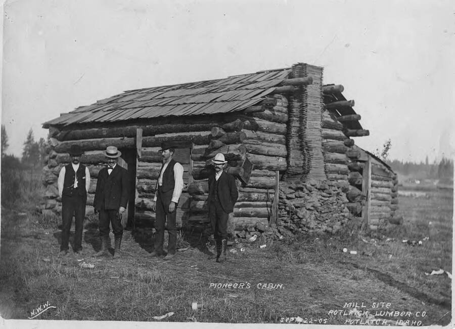 Four men stand in front of a log cabin. According to what is written on the photograph this is 'pioneer's cabin' and future 'mill site'. Written on the back of the photograph says 'man in straw hat looks like Mr. C. N Lirekenson who built the mill ('Bud' Lirekinson's grandfather).'