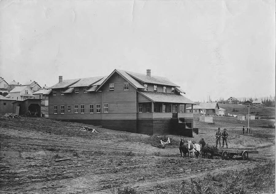 According to the description on the back of the photograph, this is the 'permanent co. boarding house.' In front of the house are two men standing on a wagon load driving a four hitch.