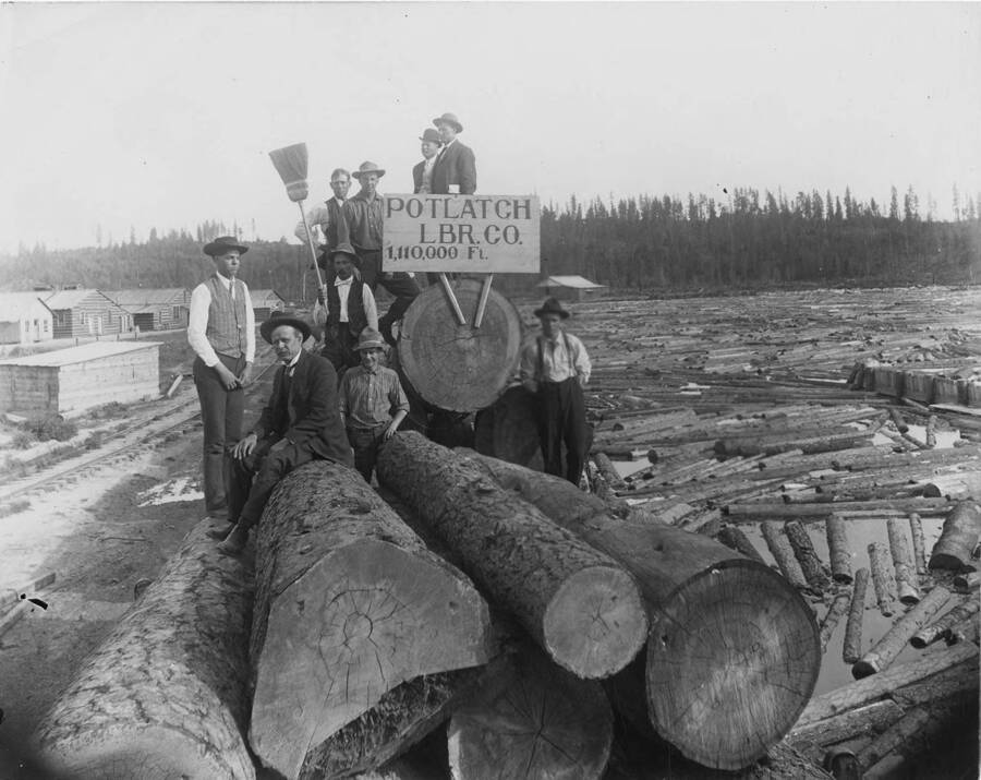 A group of Potlatch men stand and sit on top of logs, next to one of the log pods at the Potlatch mill. The sign attached to one of the logs reads 'Potlatch LBR Co. 1,110,000.
