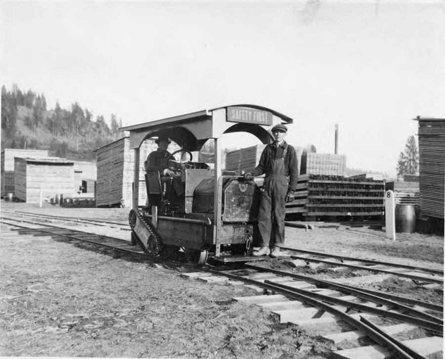 A man drives a railcar in the Potlatch mill lumber yard while another man rides on the front. Behind them are stacks of lumber.