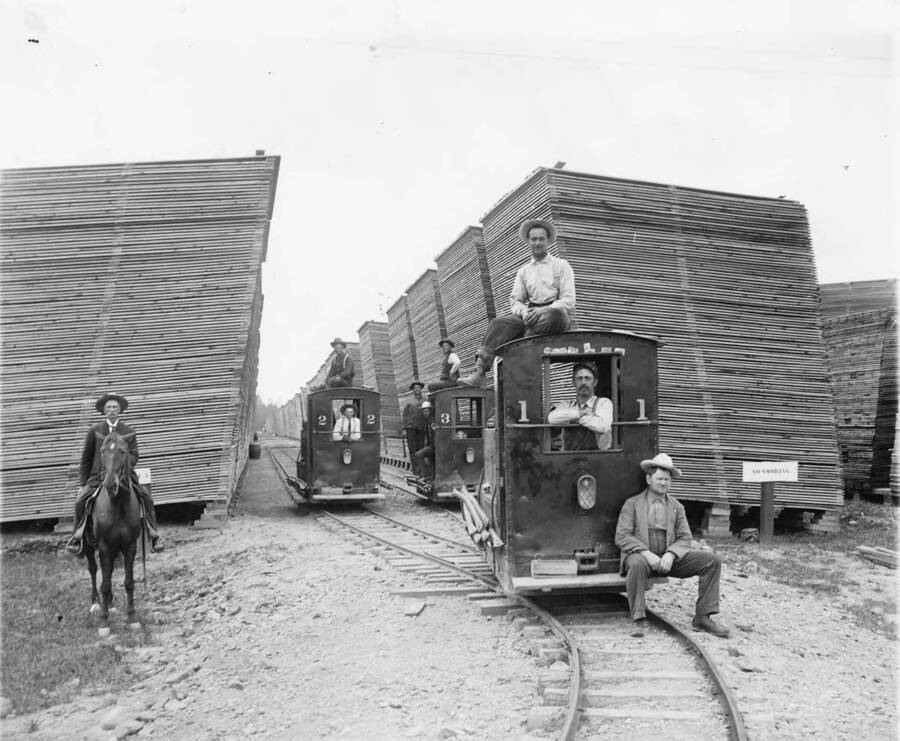 Men pose for a photograph with the cars that make up the rail system in the Potlatch lumber yard. Behind them are stacks of board lumber. One man sits on his horse on the left side of the photograph.