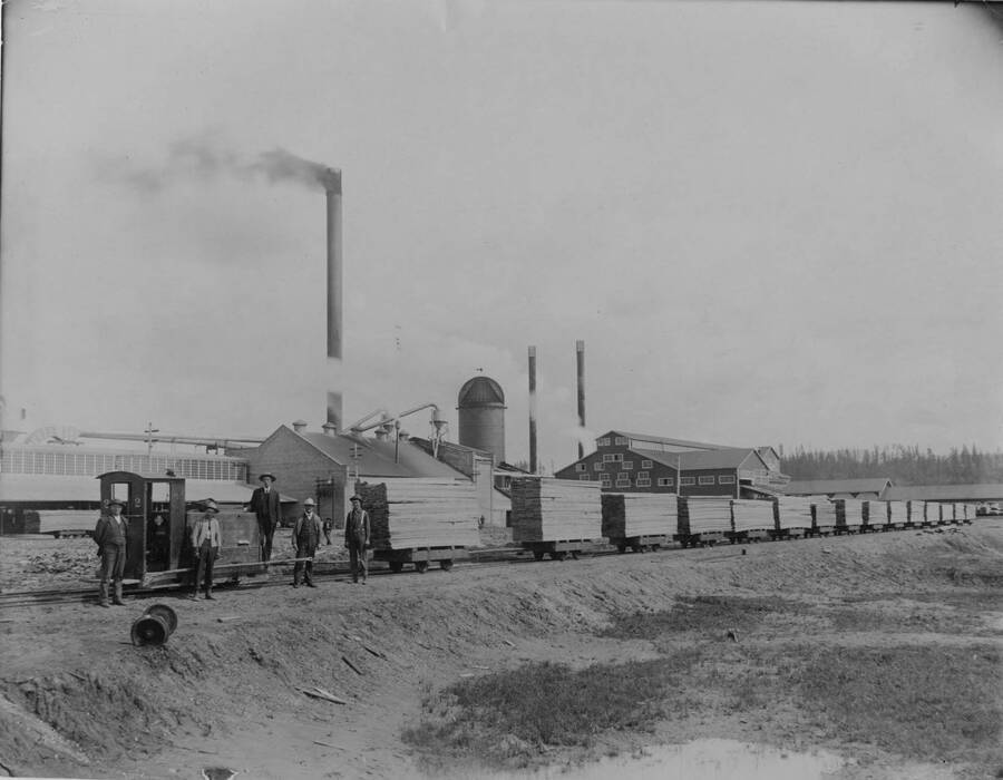 Five men stand with long train of flatcars full of lumber at the Potlatch mill which stands behind them.